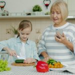 Grandmother with smartphone is teaching granddaughter to cook salad at home, cute little girl is cutting cucumber using knife and chopping board.