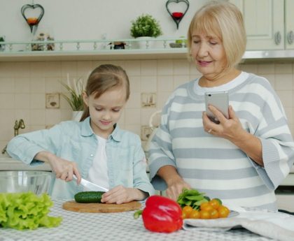 Grandmother with smartphone is teaching granddaughter to cook salad at home, cute little girl is cutting cucumber using knife and chopping board.