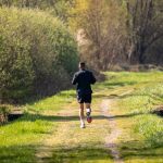 A man jogs on a scenic, grassy forest path surrounded by lush greenery under daylight.