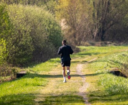 A man jogs on a scenic, grassy forest path surrounded by lush greenery under daylight.